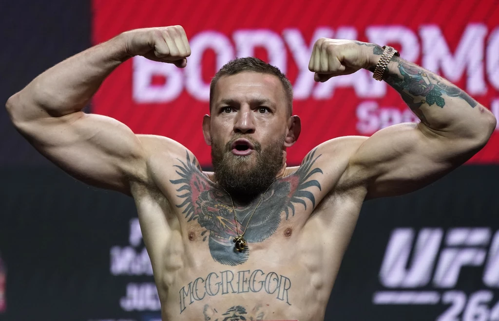 McGregor zapowiedział powrót na gali UFC w Białym Domu Mężczyzna w pozie prezentującej umięśnione ramiona i tors, z widocznymi tatuażami oraz napisem na klatce piersiowej, w tle elementy scenografii gali sportowej.
