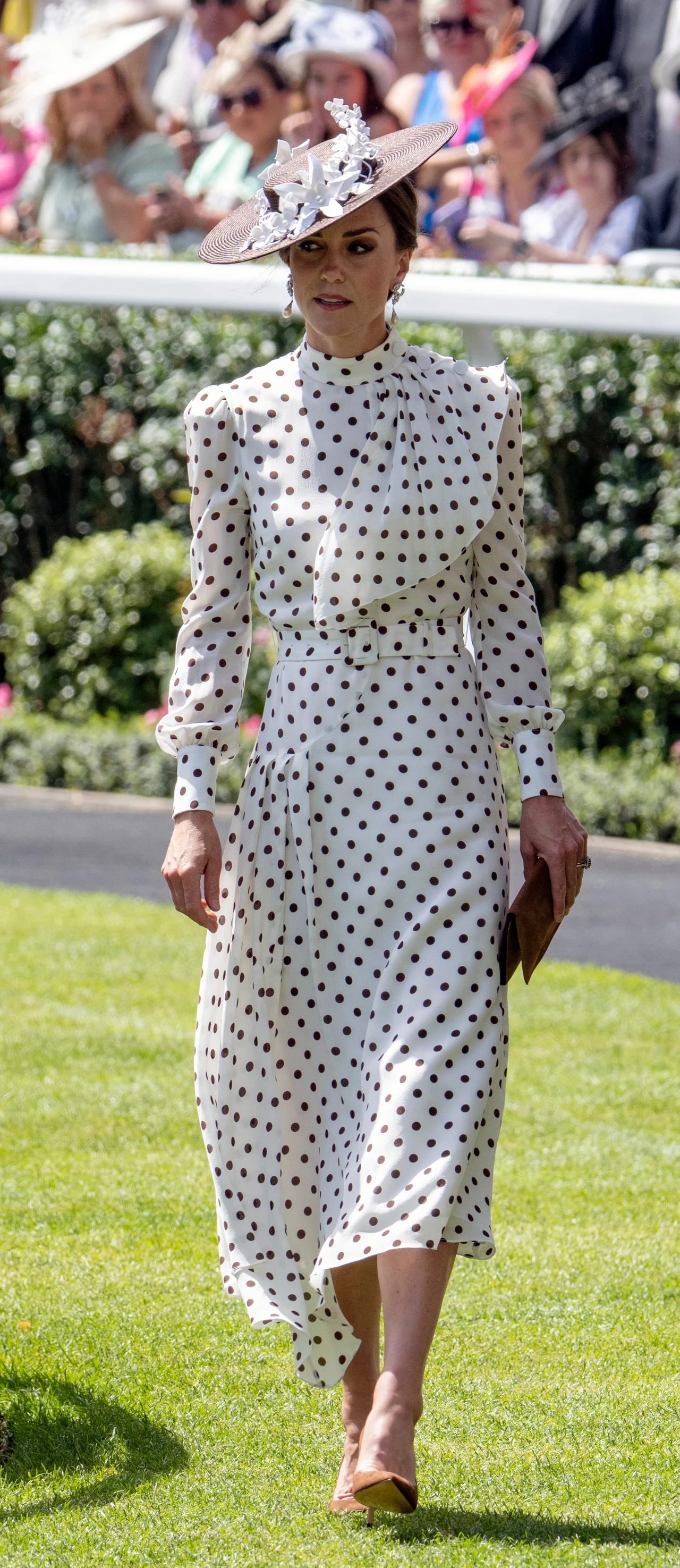 Elegancka kobieta w białej sukience w czarne grochy oraz dużym, ozdobnym kapeluszu przechadza się po trawie, trzymając w dłoni brązową kopertówkę. Za nią tłum ludzi w letnich strojach, siedzący na trybunach.