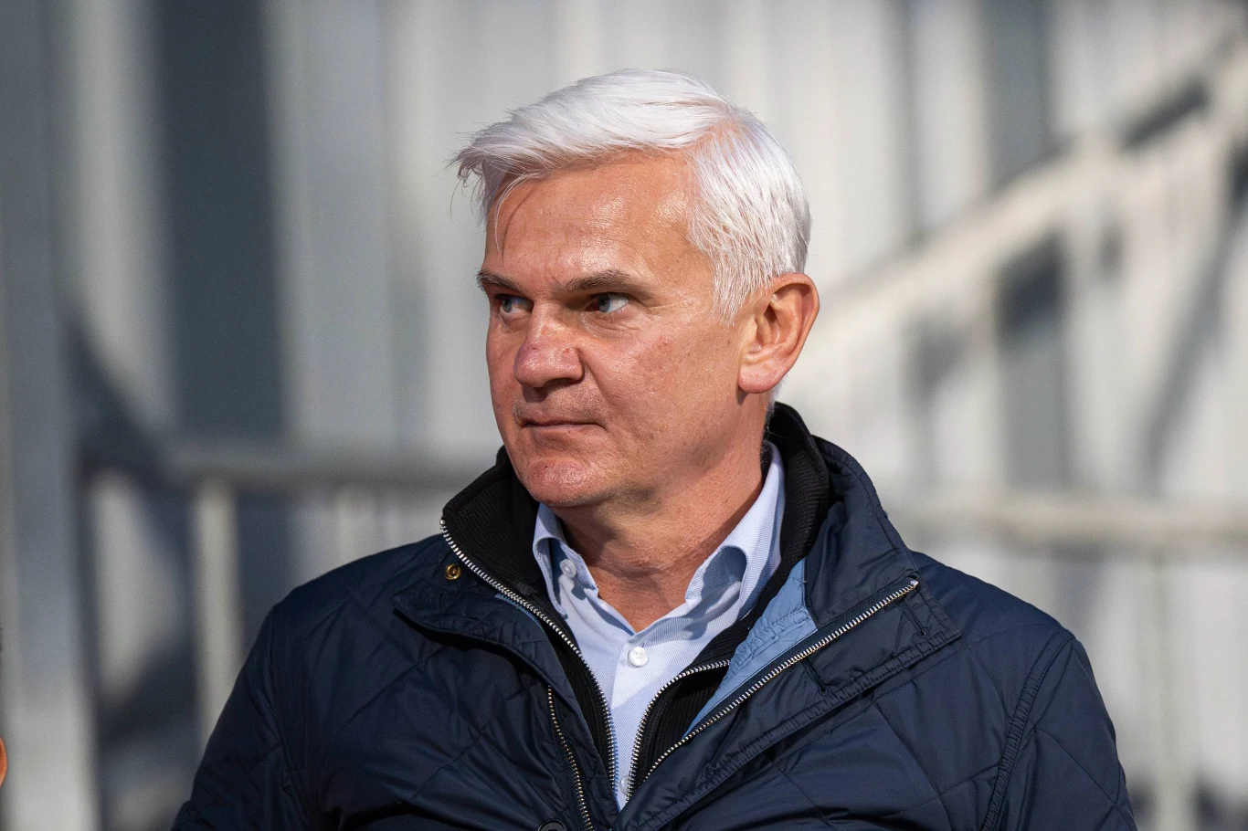 Jacek Magiera Mężczyzna z siwymi włosami w granatowej kurtce i jasnoniebieskiej koszuli patrzy w bok, stoi na tle rozmytego, jasnego budynku przemysłowego.