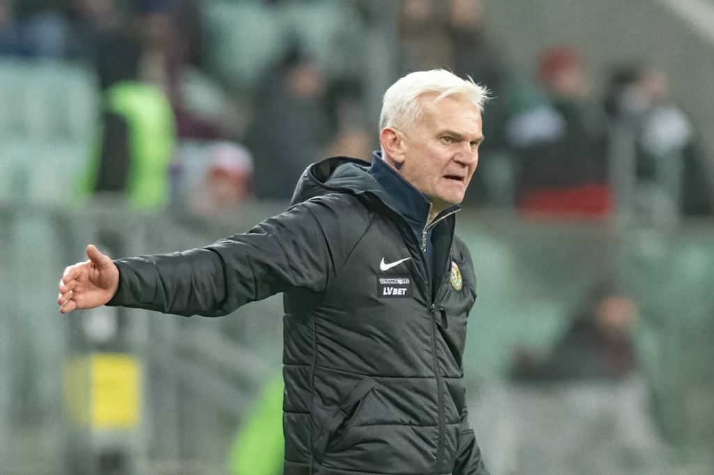 Jacek Magiera Mężczyzna z siwymi włosami w sportowej kurtce gestykuluje na tle trybun stadionu, wygląda na zaangażowanego w sytuację sportową.