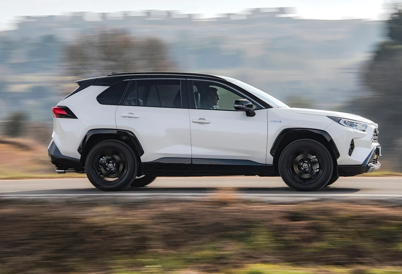 Toyota RAV4 to najlepiej sprzedające się nowe auto świata w 2024 roku Biały samochód typu SUV jedzie po asfaltowej drodze, w tle rozmyte krajobrazy i delikatnie pofałdowane wzgórza, pojazd ma czarne felgi i przyciemnione szyby.