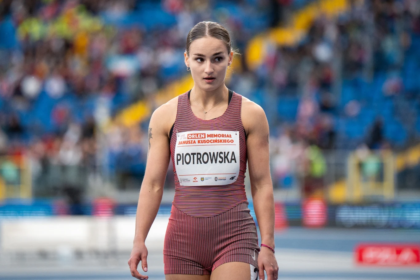 Lekkoatletka w sportowym stroju startowym stoi na stadionie lekkoatletycznym, skupiona i gotowa do rozpoczęcia biegu, w tle rozmyte trybuny z kibicami.