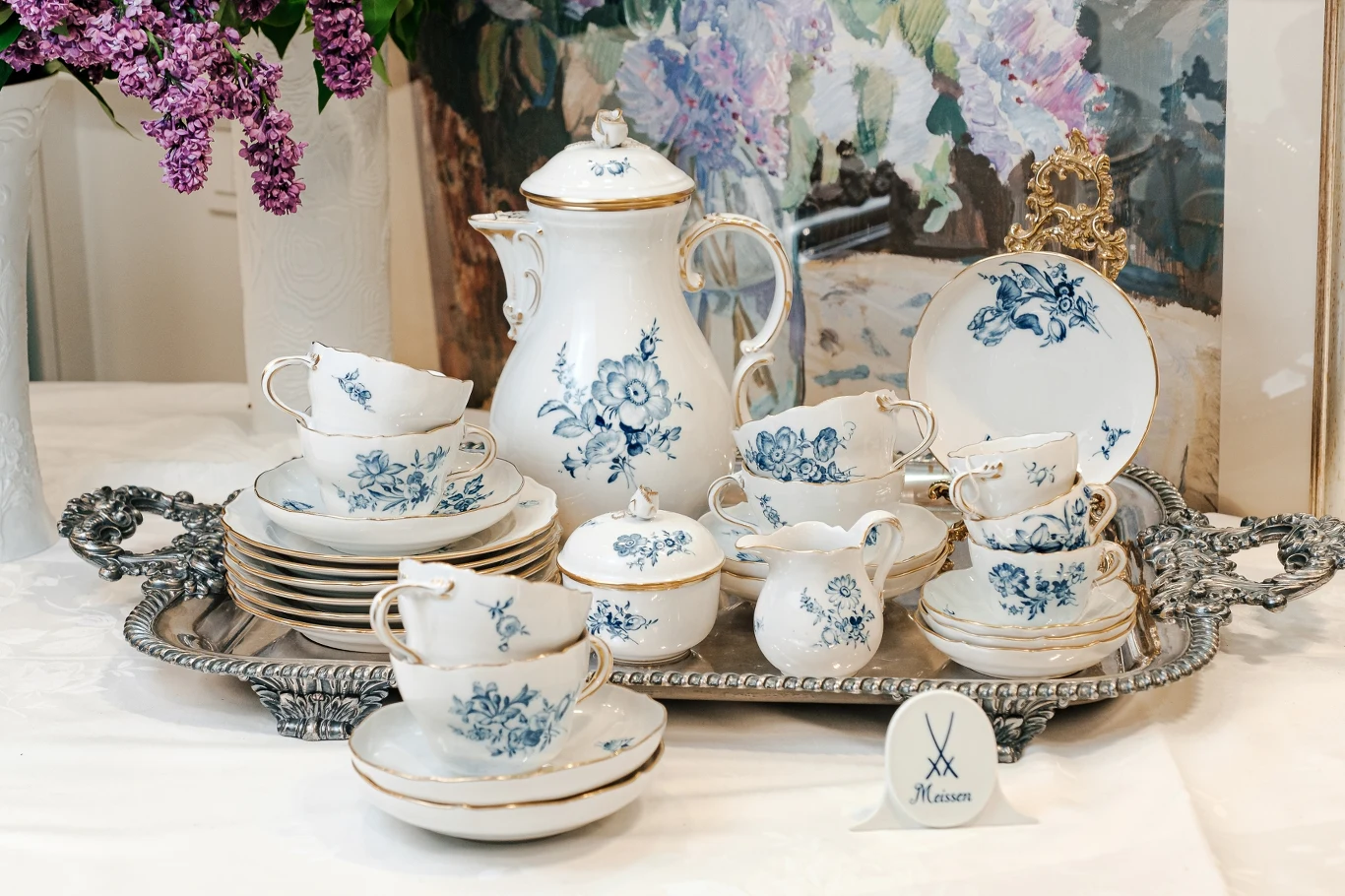 Porcelana to ponadczasowy wybór Elegancki porcelanowy serwis do herbaty zdobiony niebieskimi kwiatami, ustawiony na srebrnej tacy, otoczony przez bukiet bzu oraz obraz w tle.