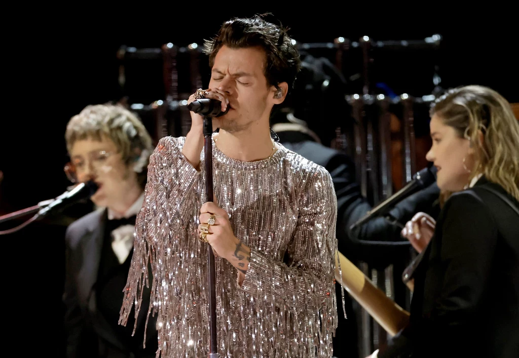 Harry Styles Wokalista ubrany w błyszczącą koszulę z frędzlami śpiewa do mikrofonu, wokół niego znajdują się inni muzycy grający na instrumentach na ciemnym tle.