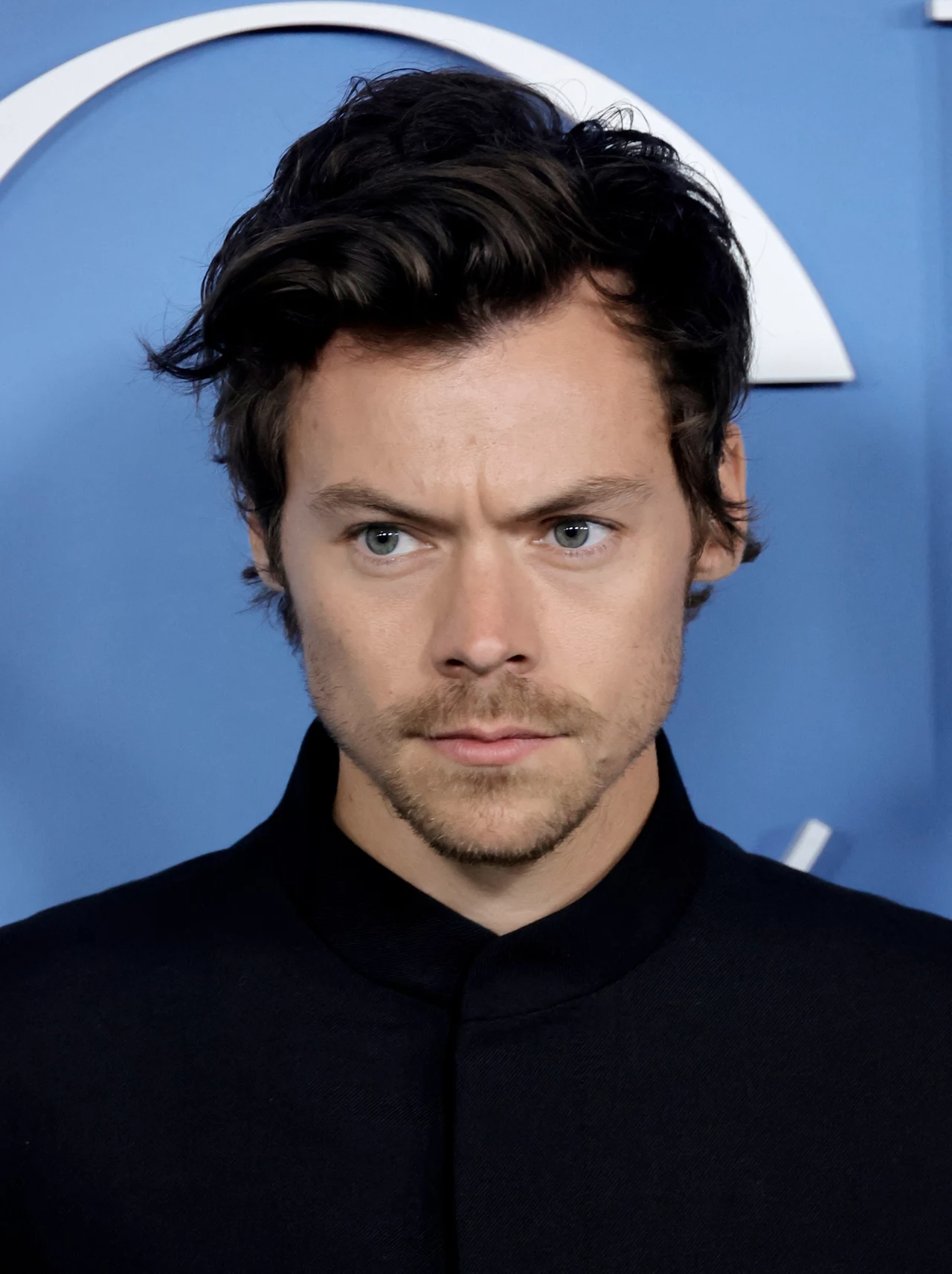 Harry Styles Mężczyzna o ciemnych włosach, ubrany w czarną marynarkę, z poważnym wyrazem twarzy na tle niebieskiej ścianki.