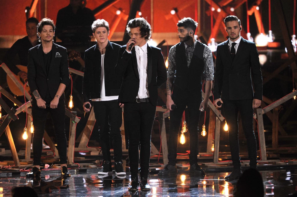 One Direction. Od lewej: Louis Tomlinson, Niall Horan, Harry Styles, Zayn Malik i Liam Payne Pięciu młodych mężczyzn ubranych elegancko, stojących na scenie wśród świateł i drewnianej scenografii, jeden z nich trzyma mikrofon i śpiewa, pozostali stoją w rzędzie z poważnym wyrazem twarzy.