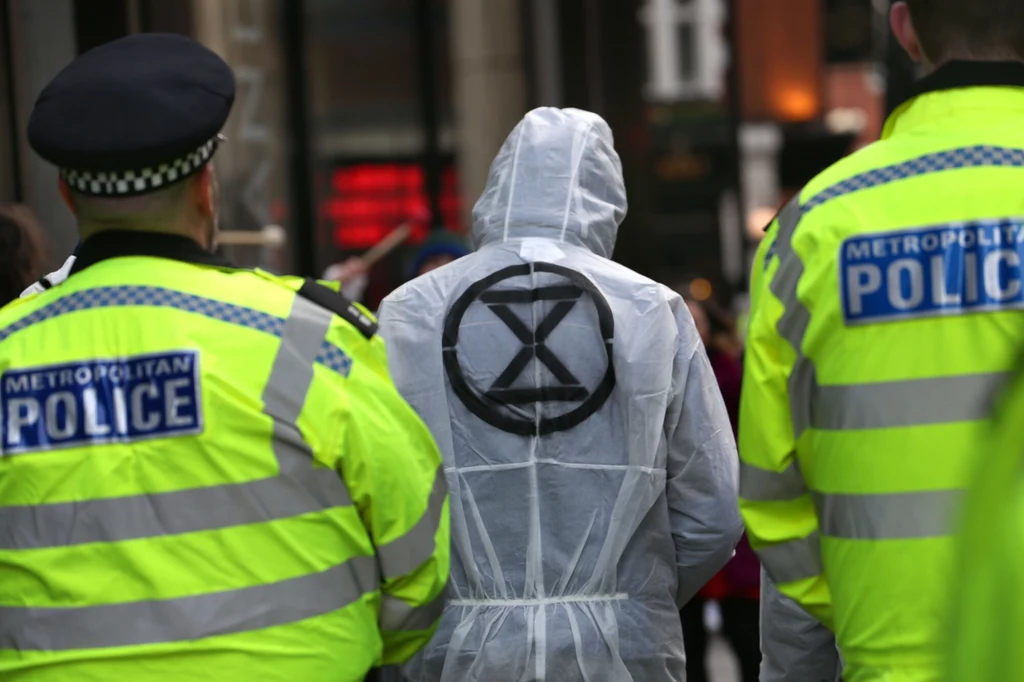 Dwóch funkcjonariuszy policji w kamizelkach odblaskowych eskortuje osobę w białym kombinezonie ochronnym z symbolem Extinction Rebellion na plecach, tło stanowią zabudowania miejskie.