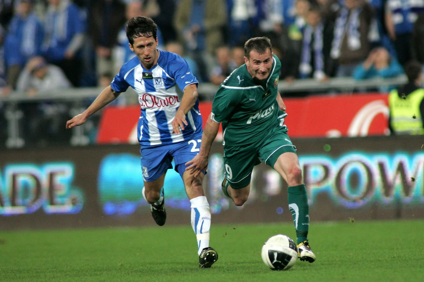 Dwóch piłkarzy podczas dynamicznej walki o piłkę na murawie, jeden z zawodników ubrany w zielony strój prowadzi futbolówkę, a drugi w niebiesko-białym stroju stara się mu ją odebrać, w tle rozmazana publiczność na trybunach.