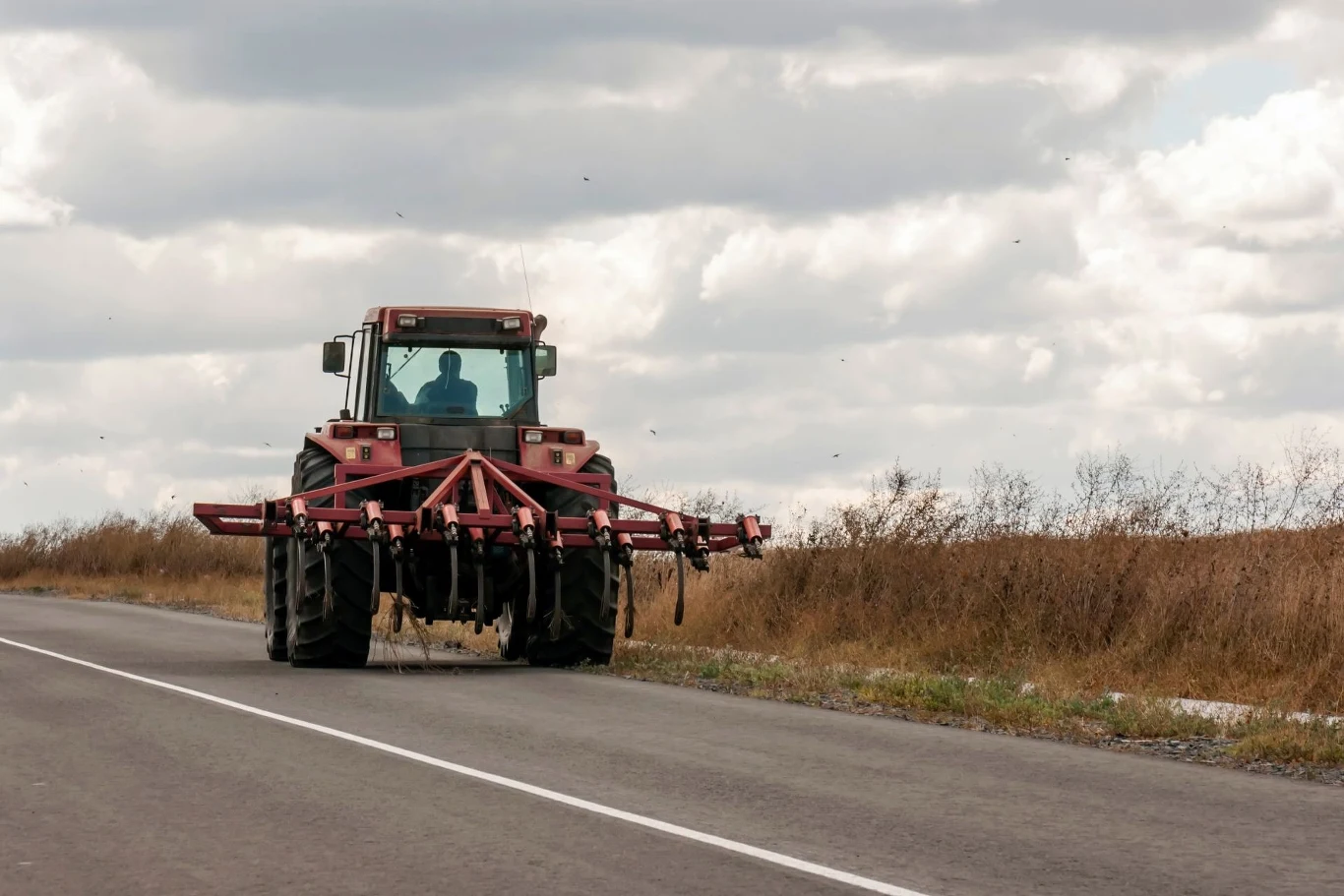 Czerwony ciągnik rolniczy jedzie samotnie asfaltową drogą poza miastem, w tle widać suche trawy i chmury na niebie.