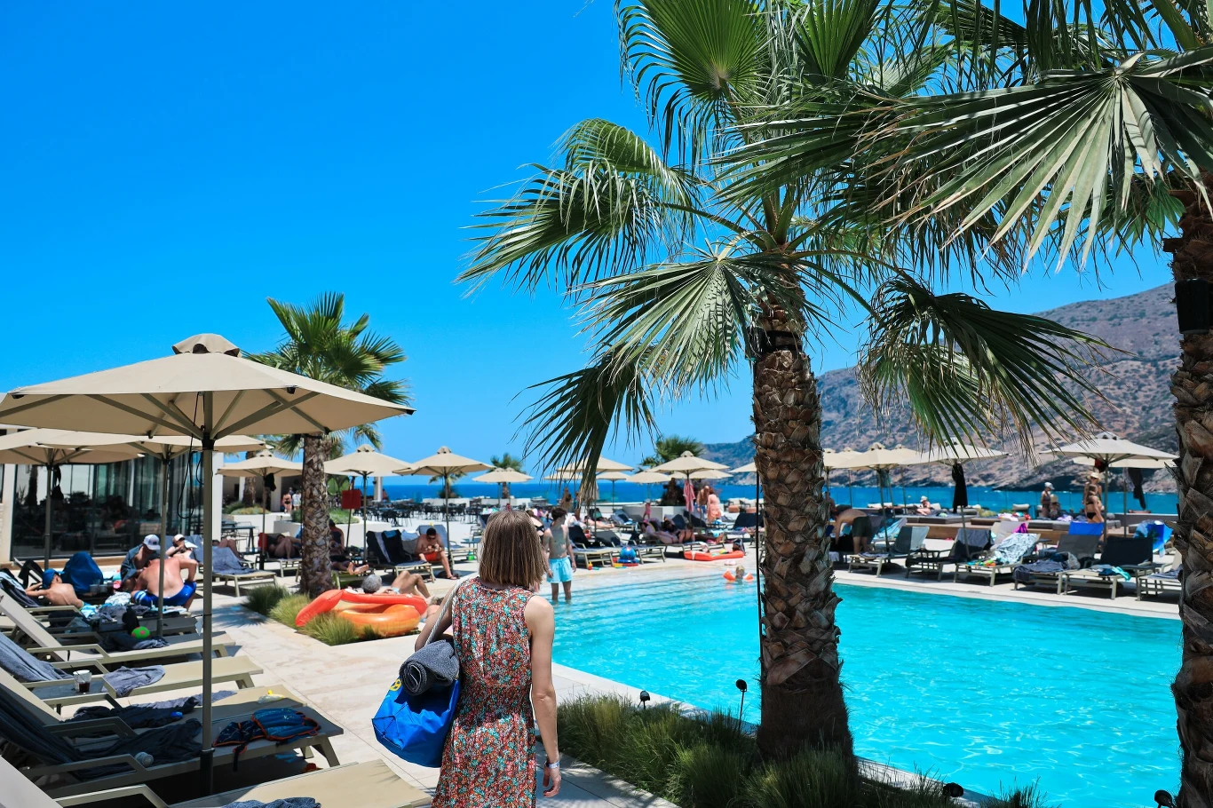 Hotele z all inclusive wprowadzają zmiany dla turystów? Basen hotelowy otoczony leżakami i parasolami, liczne osoby odpoczywające przy wodzie, w tle palmowe drzewa i widok na góry, kobieta z torbą kieruje się w stronę basenu.