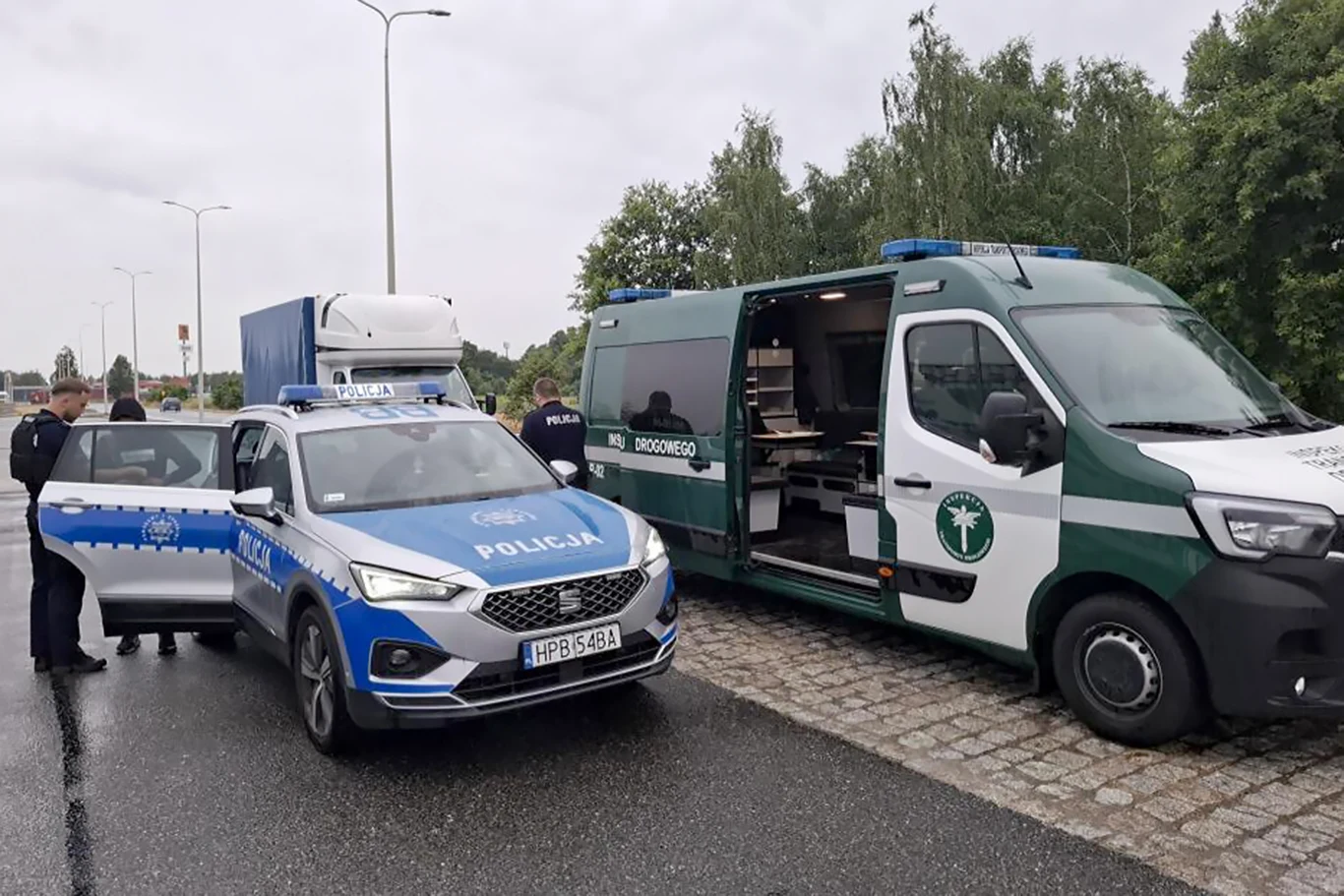 Kontrole ITD przy granicy. Usterki techniczne i fałszywe prawa jazdy | Fot. WITD Wrocław Policjanci oraz funkcjonariusze inspekcji transportu drogowego stoją obok policyjnego radiowozu i pojazdu kontrolnego ITD zaparkowanych na poboczu drogi, w tle widoczna ciężarówka i drzewa.