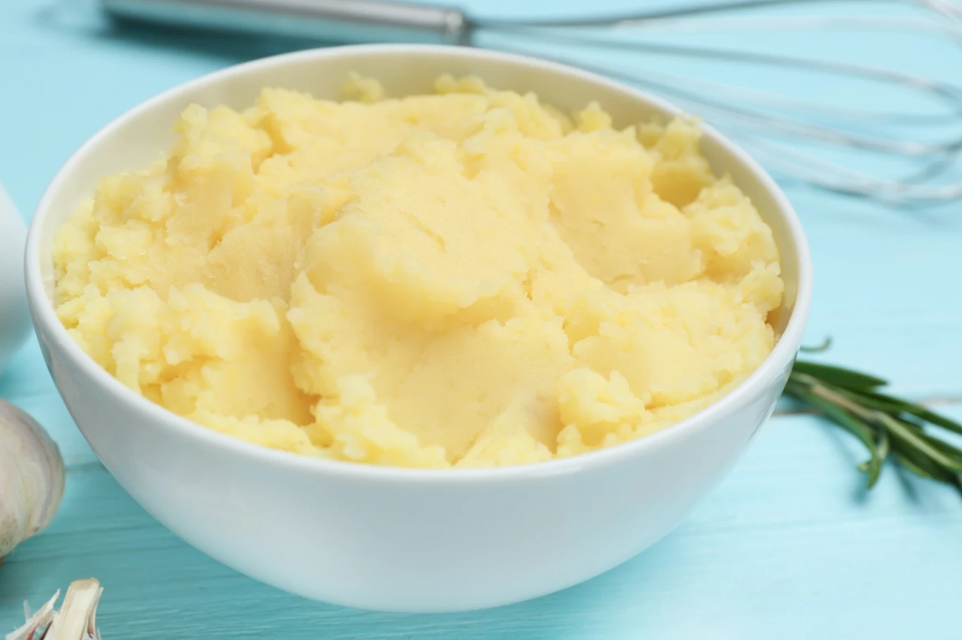 Gładkie puree ziemniaczane o kremowej konsystencji w białej misce, na niebieskim blacie z widocznym z boku ząbkiem czosnku, gałązką rozmarynu oraz fragmentem trzepaczki kuchennej.