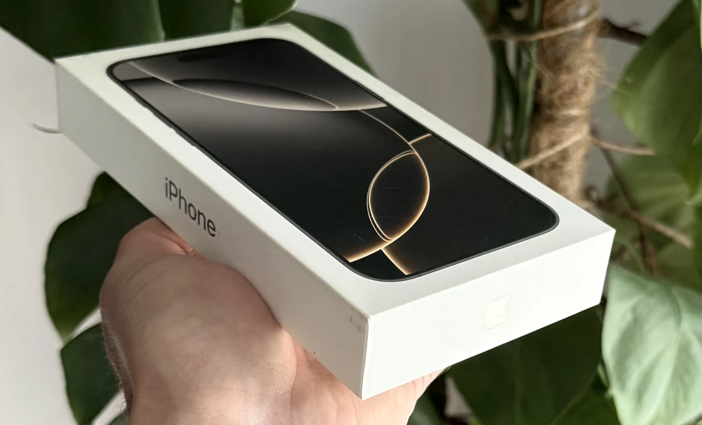 Nowy iPhone nie każdego roku? Apple zmienia strategię. Nowy telefon iPhone w białym opakowaniu trzymany w dłoni na tle zielonych liści rośliny.