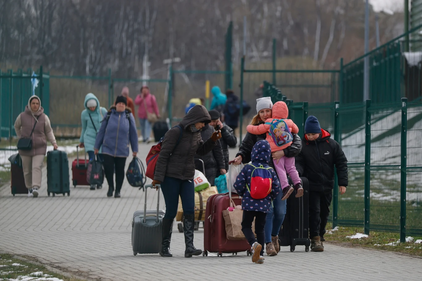 Ukraińcy przekraczający polską granicę w 2022 Ludzie idący chodnikiem, niosący walizki i plecaki, niektórzy prowadzą dzieci za rękę lub niosą je na rękach, w tle metalowe ogrodzenie i pora zimowa, widoczne resztki śniegu.