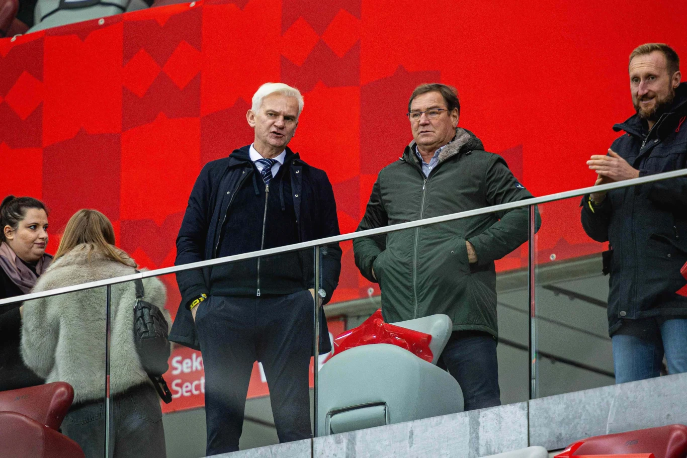 Jacek Magiera i Jan Urban Grupa osób w eleganckich płaszczach i kurtkach stoi na trybunach stadionu, na tle dużego czerwonego baneru, obejmują rozmowę i obserwację wydarzeń sportowych.