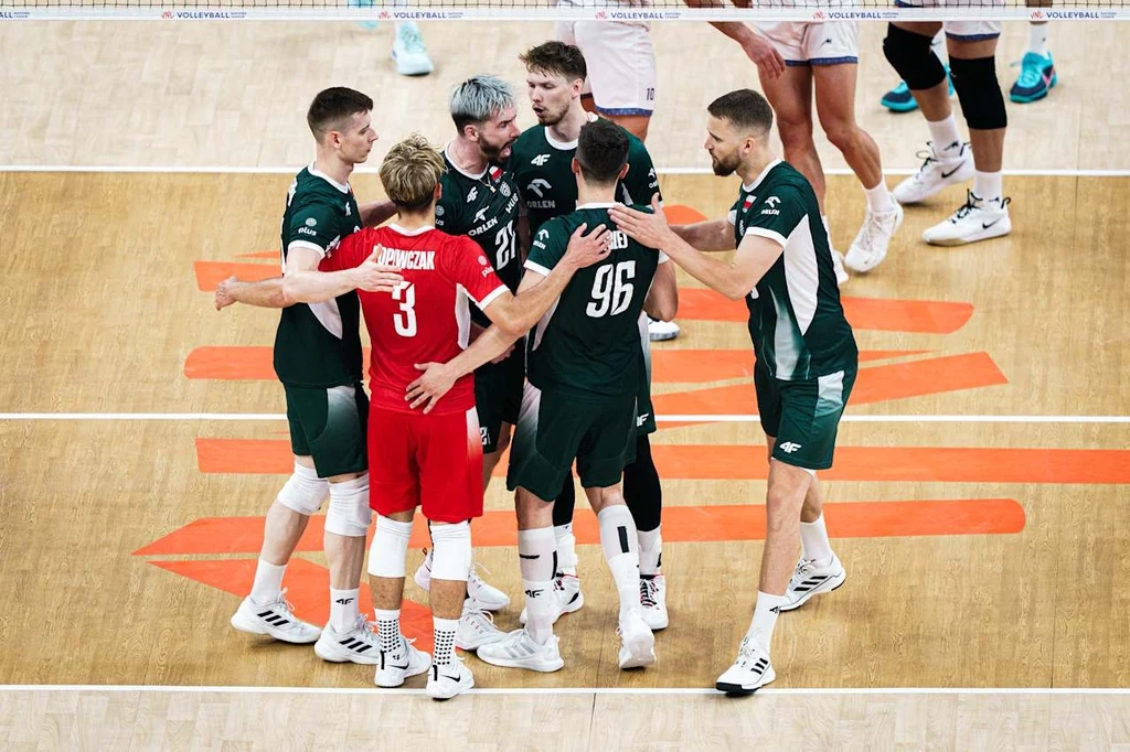 Liga Narodów siatkarzy. Jakub Popiwczak (w czerwonej koszulce) i koledzy z reprezentacji Polski Grupa zawodników drużyny siatkarskiej ubranych w zielone i czerwone stroje zgromadzona na środku boiska, świętująca zdobyty punkt podczas meczu, widoczne emocje i współpraca zespołowa.