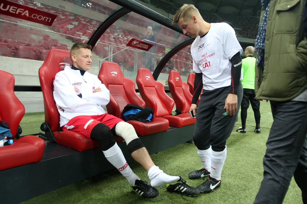 Marcin Mroczek i Rafał Mroczek Dwóch piłkarzy w sportowych strojach rozmawia na ławce rezerwowych na stadionie, jeden z nich siedzi z opatrunkami na kolanach, drugi stoi obok, atmosfera sugeruje przerwę lub oczekiwanie na wejście do gry.