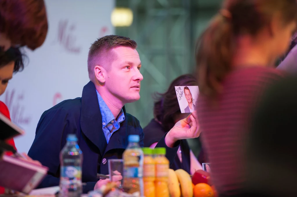 Marcin Mroczek Mężczyzna siedzi przy stole, trzyma w ręce fotografię z portretem, wokół niego znajdują się inne osoby oraz różnorodne produkty spożywcze takie jak owoce i butelki z napojami.