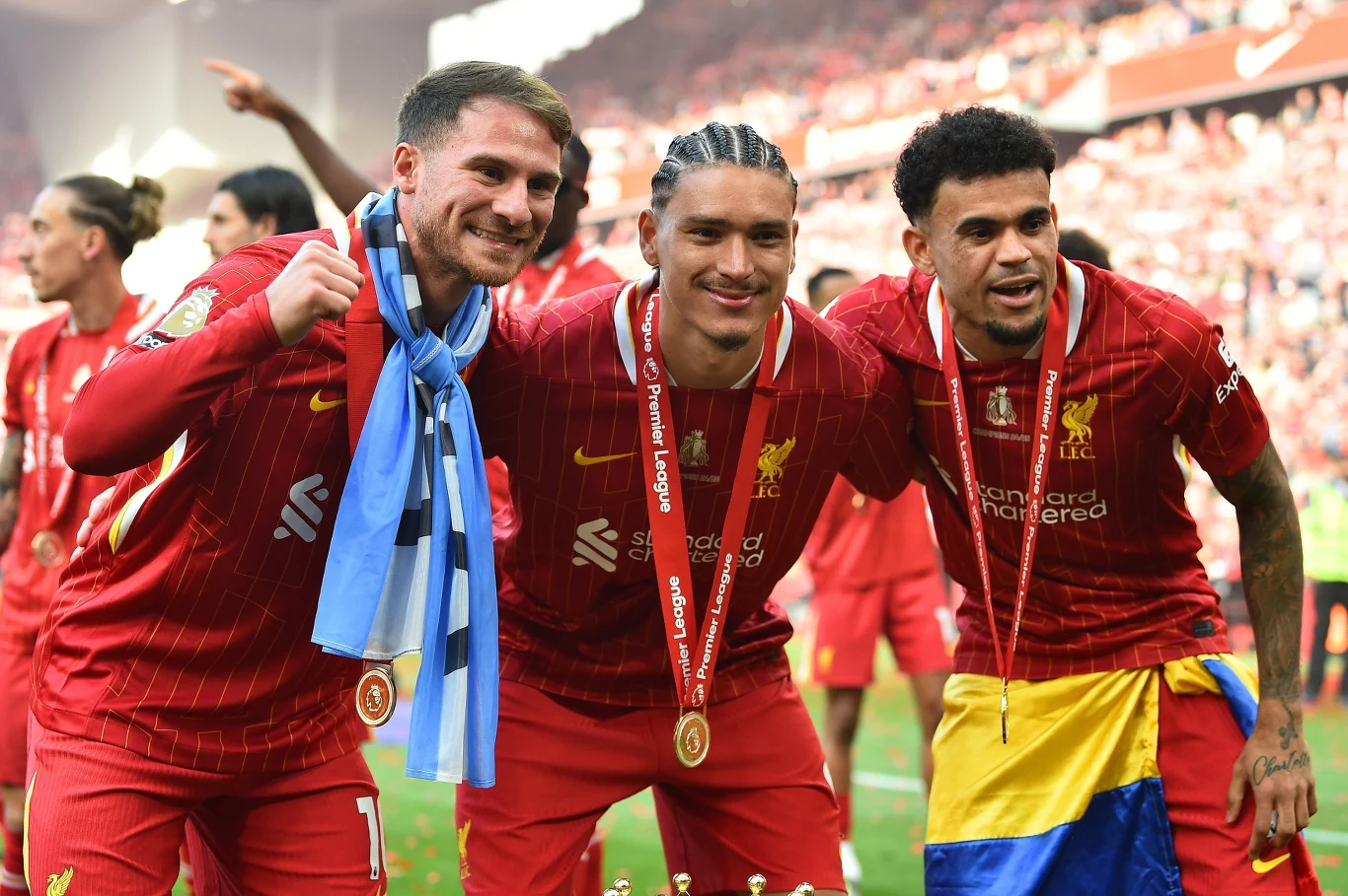 Alexis Mac Allister, Darwin Nunez i Luis Diaz Trzech piłkarzy drużyny Liverpool ubranych w czerwone stroje świętuje na stadionie, mają medale na szyjach i szerokie uśmiechy na twarzach, jeden z zawodników ma przewieszoną przez ramiona niebiesko-błękitną flagę, a inny owinięty jest żółto-niebieską ...