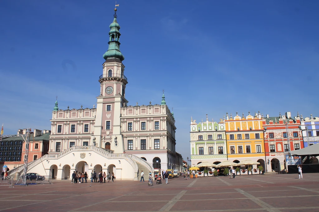 Renesansowy ratusz z wysoką wieżą na środku dużego, otwartego rynku, wokół kolorowe kamienice charakteryzujące się bogatą ornamentyką fasad, na placu widoczne są grupy spacerujących ludzi.