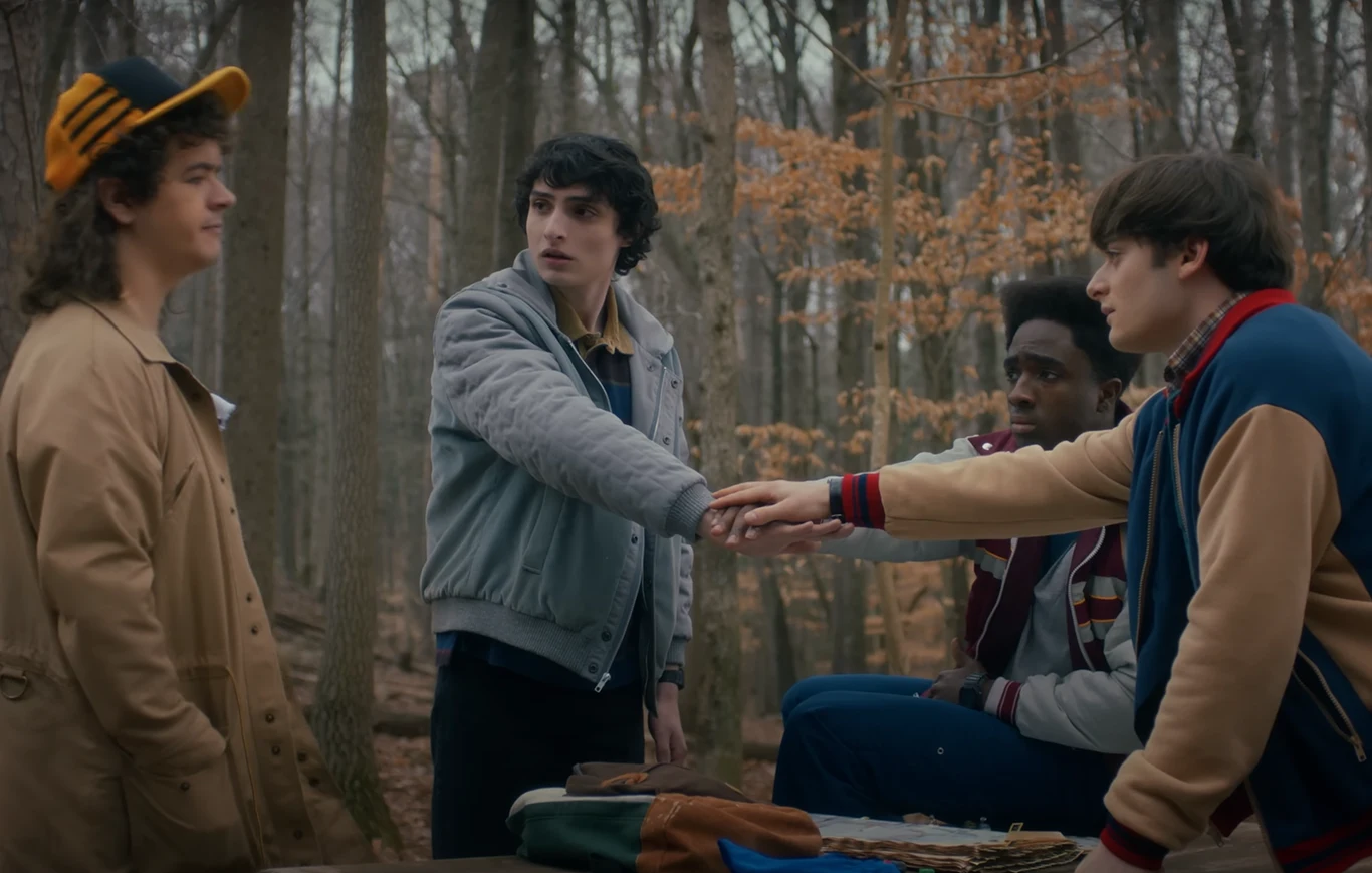 Kadr ze zwiastuna "Stranger Things 5" Czterech nastoletnich chłopców trzyma ręce razem nad stołem na tle jesiennego lasu, każdy z nich skupiony na wspólnym geście, ubrani w casualowe, kolorowe kurtki i swetry, wokół rozłożone są gry planszowe.