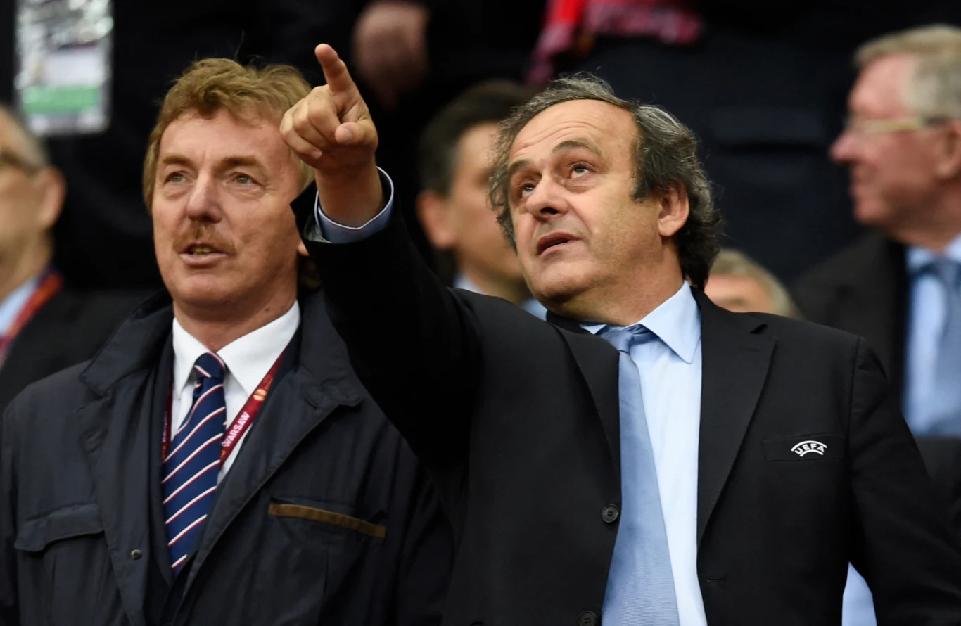 Michel Platini (z prawej) w towarzystwie swojego przyjaciela, Zbigniewa Bońka Dwóch elegancko ubranych mężczyzn w garniturach, jeden z nich energicznie wskazuje coś w oddali gestem ręki, obaj zaangażowani w obserwację, w tle zamazana sylwetka innych osób.