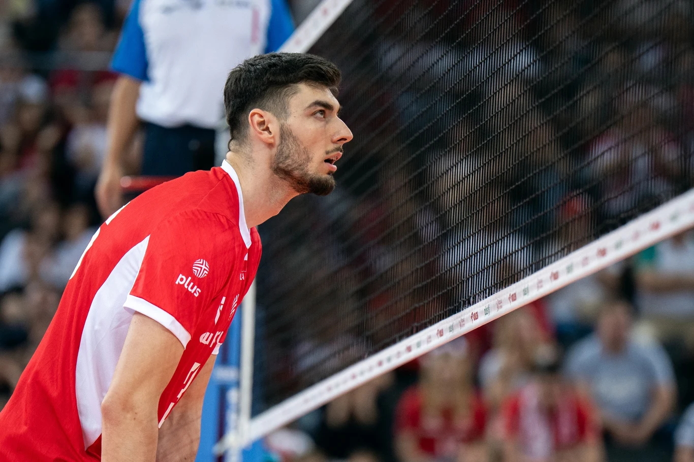 Kewin Sasak Siatkarz w czerwonym stroju stoi przy siatce w trakcie meczu, skupiony i gotowy do gry, w tle widoczni kibice na trybunach.