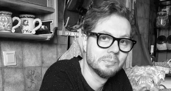 Nie żyje Rafał Zapała Mężczyzna w okularach siedzi w kuchni, w tle widoczne są dekoracyjne kubki i półki, obok towarzyszy mu niewielki pies.