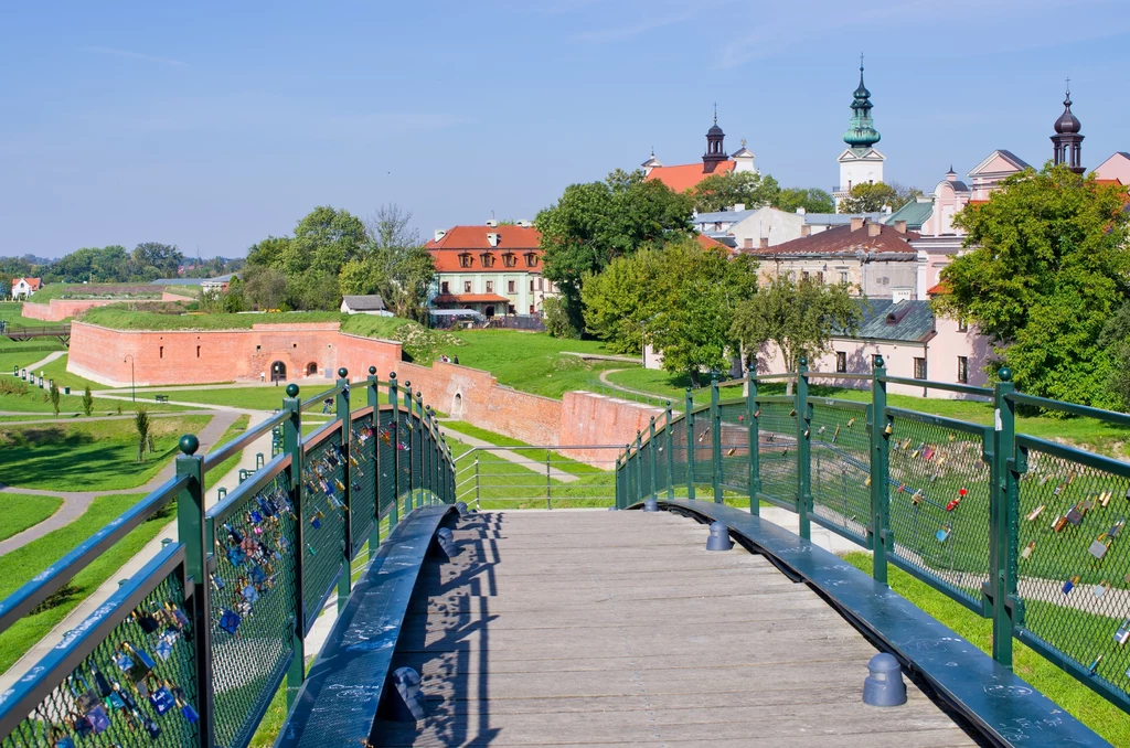 Widok z mostku z zawieszonymi kłódkami zakochanych na zielone tereny wokół ceglanych murów obronnych i historyczną zabudowę z kościołami i kamienicami w tle, całość w słoneczny dzień.