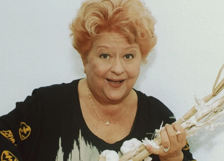 Danuta Rinn Kobieta o rudych włosach z szerokim uśmiechem trzyma w ręku pęk czosnku, ubrana w ciemną bluzkę z delikatnym wzorem, stoi na jasnym tle.