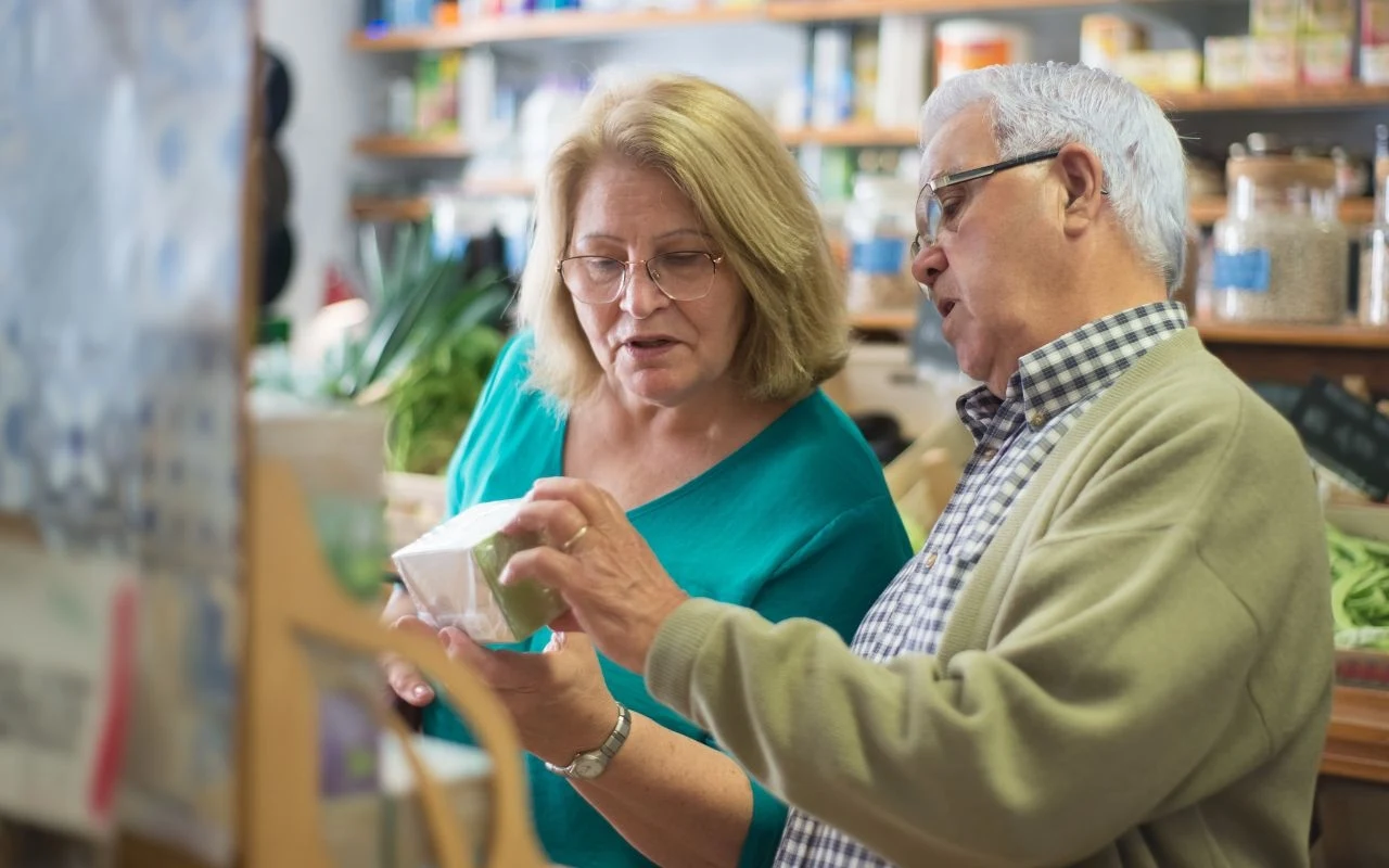 Już niebawem dla klientów wszystko stanie się jasne. Koniec z małymi, nieczytelnym oznaczeniami Starsza kobieta i starszy mężczyzna wspólnie oglądają opakowanie produktu w sklepie, analizując etykietę lub skład, półki w tle są wypełnione różnymi towarami.