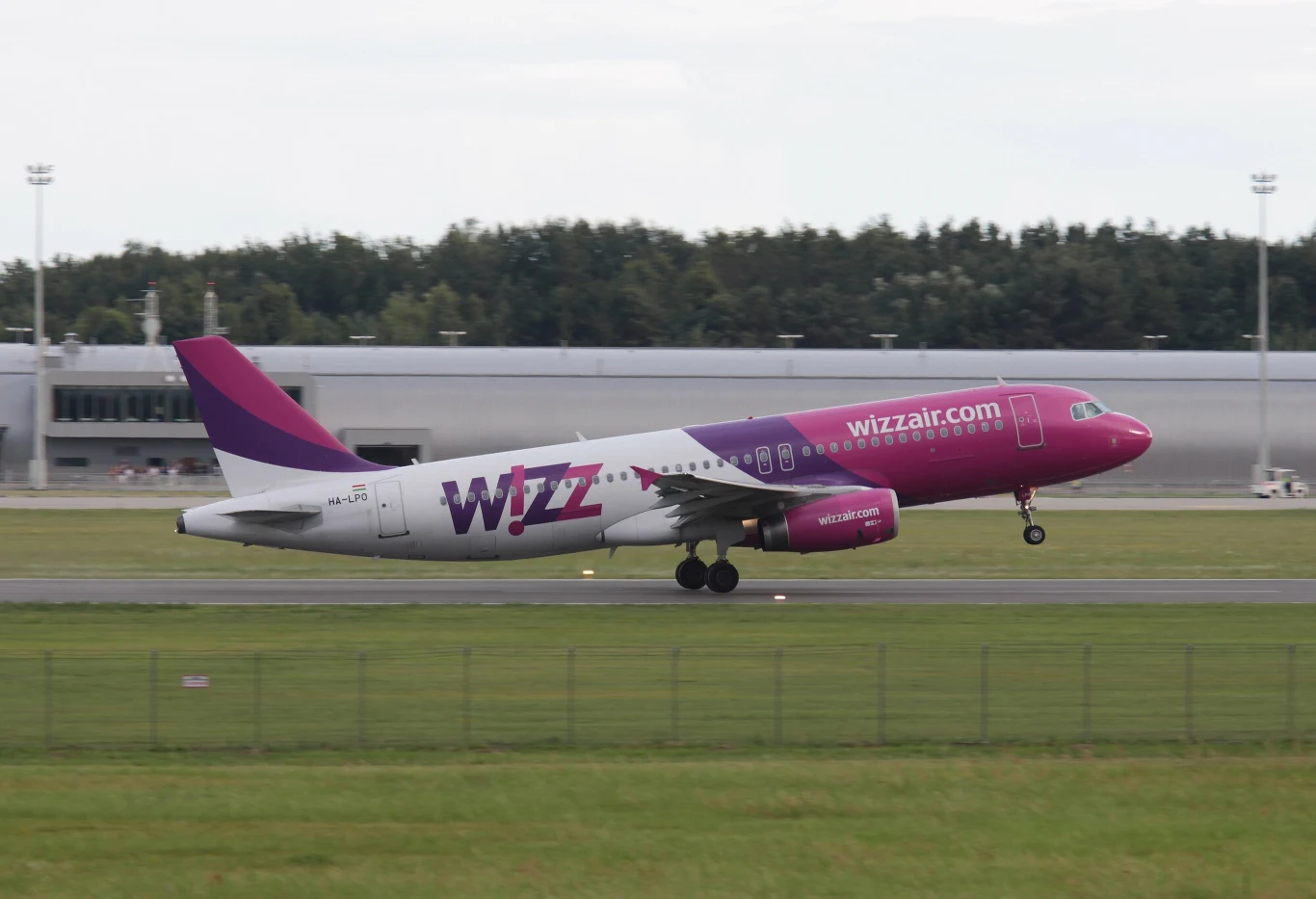 Wizz Air zimą wraca na lotnisko Warszawa-Modlin Samolot pasażerski linii Wizz Air w charakterystycznych barwach fioletowo-różowych ląduje na pasie startowym lotniska, w tle widoczny budynek terminala oraz zieleń drzew.