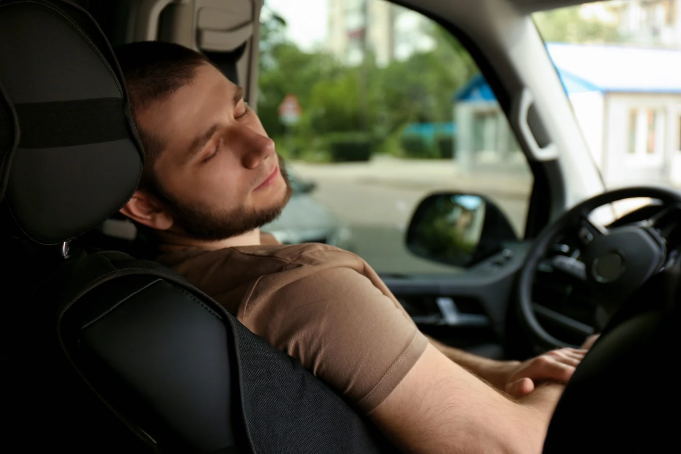 Spanie w samochodzie może skończyć się mandatem Młody mężczyzna drzemiący na siedzeniu kierowcy w zaparkowanym samochodzie, z opuszczoną głową i zamkniętymi oczami, wnętrze auta wyraźnie widoczne, w tle rozmazany obraz miejskiego otoczenia.