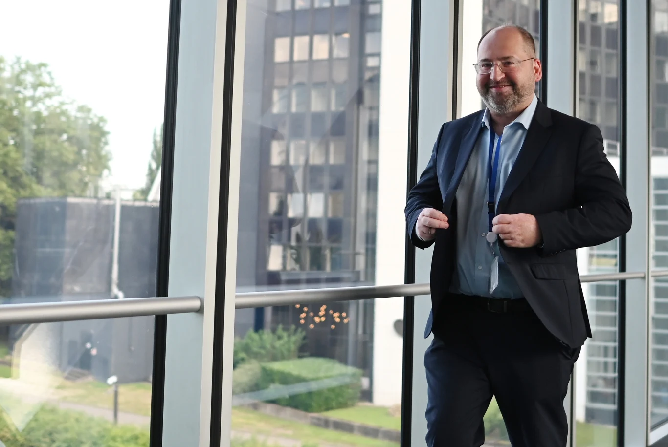 Adam Bielan broni Szymona Hołowni przed krytyką ze strony koalicjantów Mężczyzna w garniturze z identyfikatorem na szyi spaceruje po przeszklonym korytarzu z widokiem na nowoczesne budynki i zieleń za oknem.