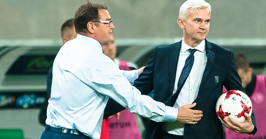 Jan Urban (z lewej) i Jacek Magiera, obecnie jego pierwszy asystent w drużynie narodowej Dwóch mężczyzn ubranych w garnitury na tle stadionu piłkarskiego, jeden z nich trzyma piłkę nożną, drugi dotyka jego brzucha w geście zatrzymania lub rozmowy, w tle ławka rezerwowych i sportowe otoczenie.