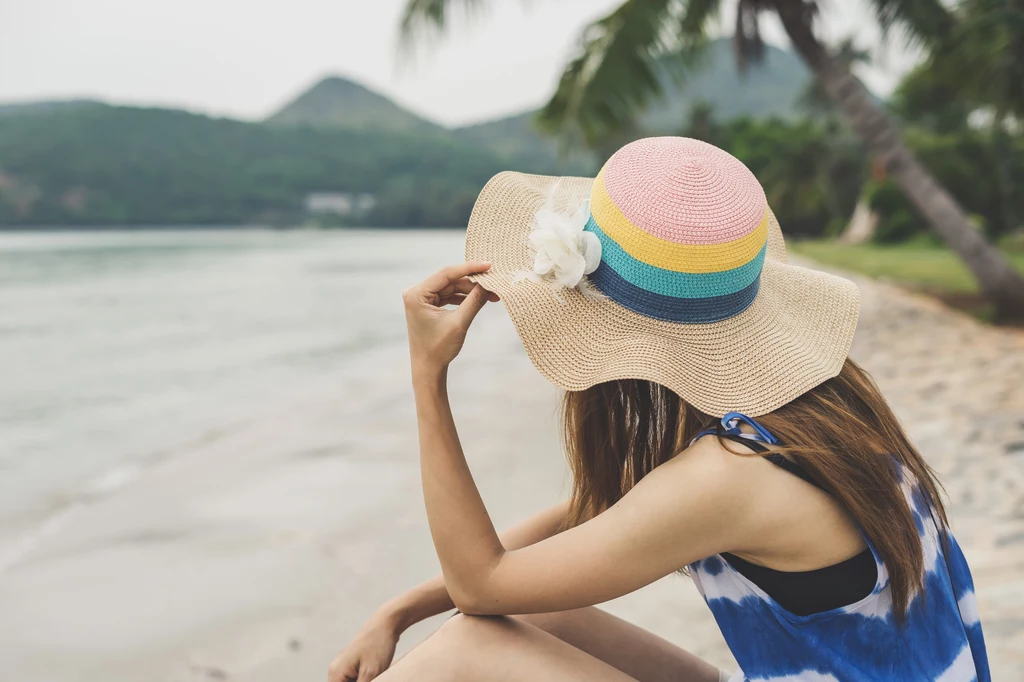 Wspólne wakacje? Tylko nie z tym znakiem zodiaku Kobieta w kolorowym kapeluszu z szerokim rondem siedzi na plaży, patrząc na morze, w tle palmy oraz wzgórza.