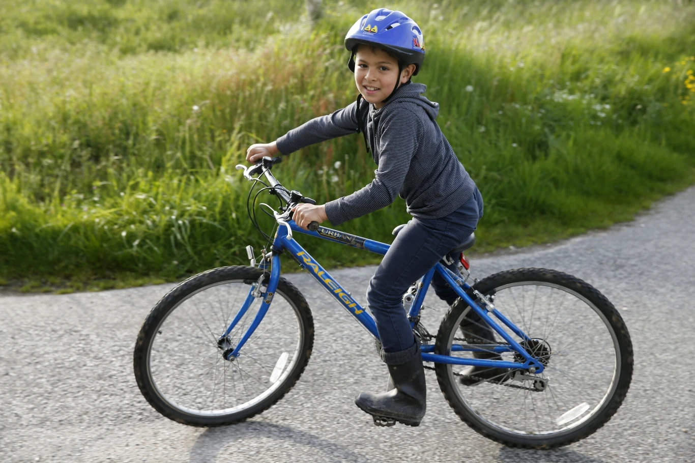 Chłopiec w kasku rowerowym jedzie niebieskim rowerem po asfaltowej ścieżce, uśmiecha się i ma na sobie ciemne ubrania oraz kalosze, w tle zielona trawa.