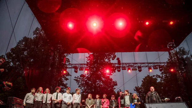 Grupa osób ustawionych w rzędzie na scenie podczas festiwalu muzycznego, oświetlonych intensywnym czerwonym światłem, w tle drzewa i nagłośnienie, na podeście napis Jarocin.