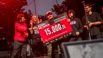 Grupa młodych osób na scenie trzyma duży symboliczny czek na 15 000 zł, otoczona światłami sceny i elementami nagłośnienia, wszyscy wyrażają radość i dumę z otrzymanej nagrody.