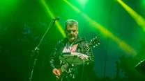 Mężczyzna grający na gitarze elektrycznej na scenie koncertowej, wokół zielone światła i dym sceniczny tworzący dynamiczną atmosferę występu na żywo.