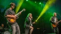 Grupa muzyków występujących na scenie podczas koncertu, na pierwszym planie gitarzysta, wokalistka śpiewająca do mikrofonu i gitarzysta po prawej stronie; w tle widoczny perkusista oraz kolorowe światła sceniczne.