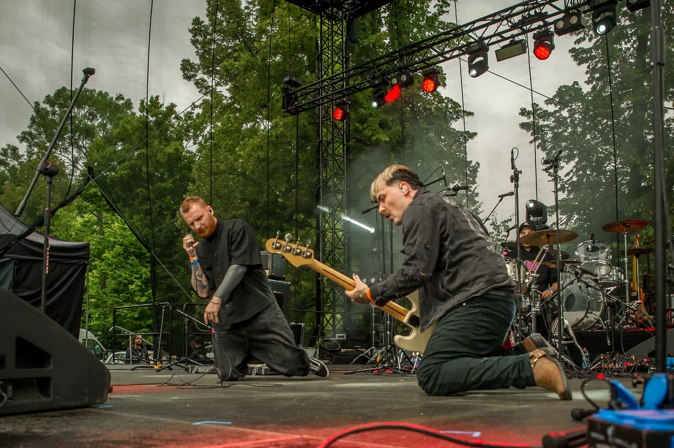 Wielkopolskie Rytmy Młodych 2025: Cafetrauma Dwóch muzyków na scenie podczas koncertu plenerowego, jeden śpiewa do mikrofonu klęcząc, drugi gra na gitarze basowej, w tle widać perkusję i drzewa, a nad sceną zawieszone są reflektory.