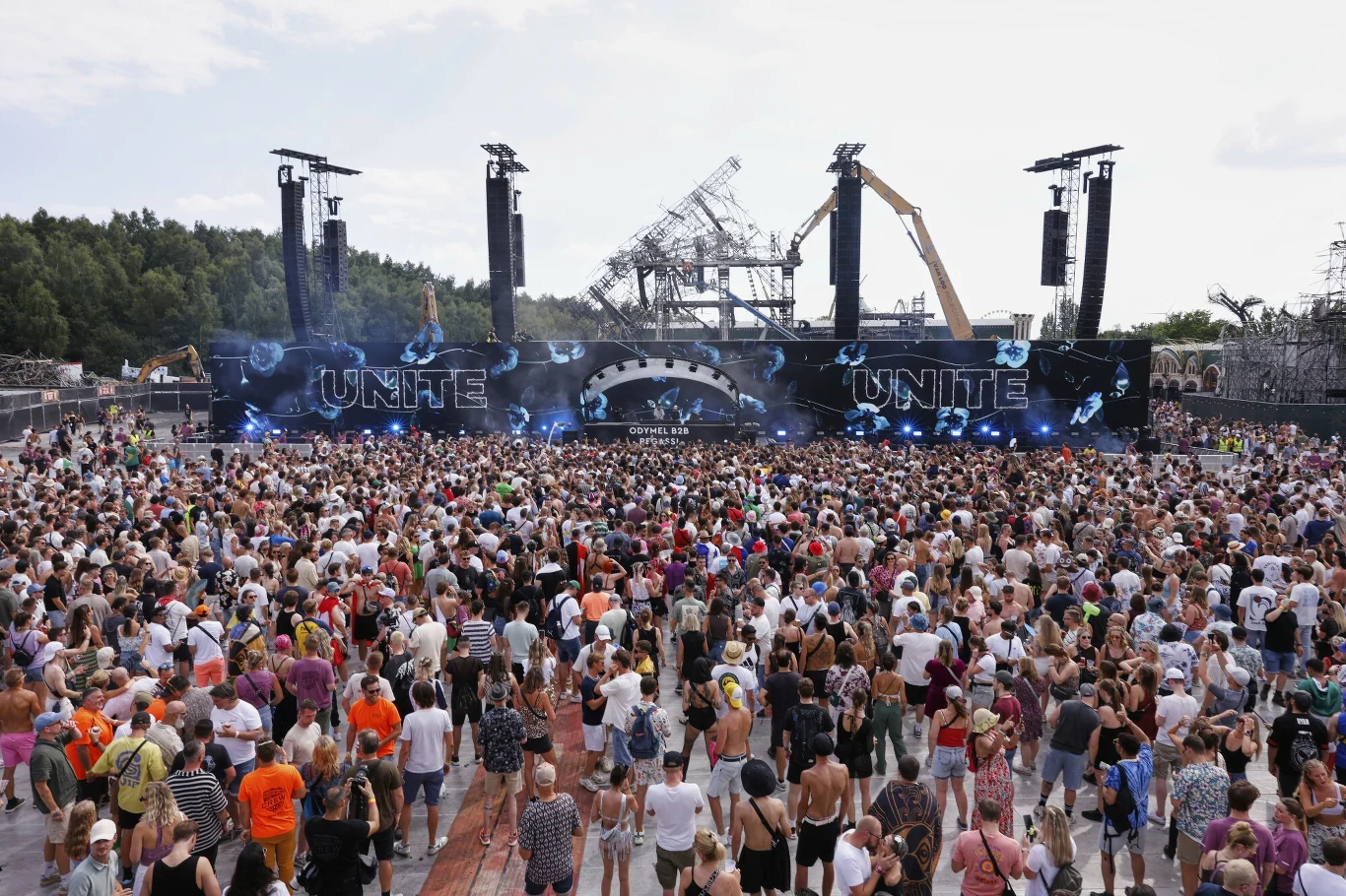 Tomorrowland 2025 wystartował, w tle resztki spalonej głównej sceny Tłum ludzi zgromadzony przed sceną koncertową z wieloma elementami świetlnymi, dużymi głośnikami i napisem UNITE na telebimach, w tle widoczna jest industrialna konstrukcja i las.