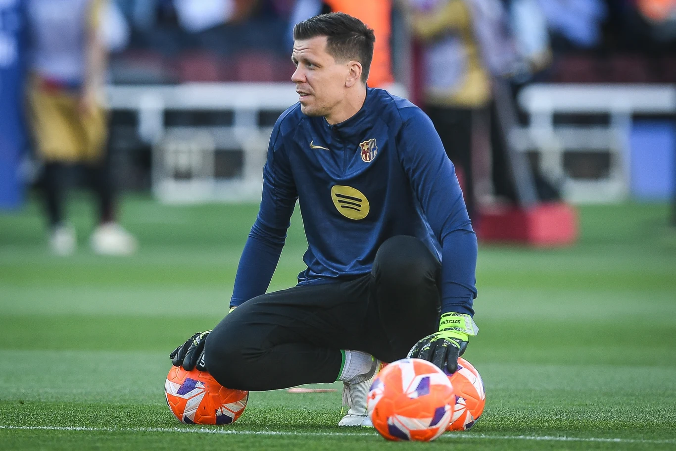 Wojciech Szczęsny Piłkarz w stroju sportowym siedzi na murawie stadionu, podpierając się rękami na trzech piłkach. W tle rozmyta trybuna z widzami.