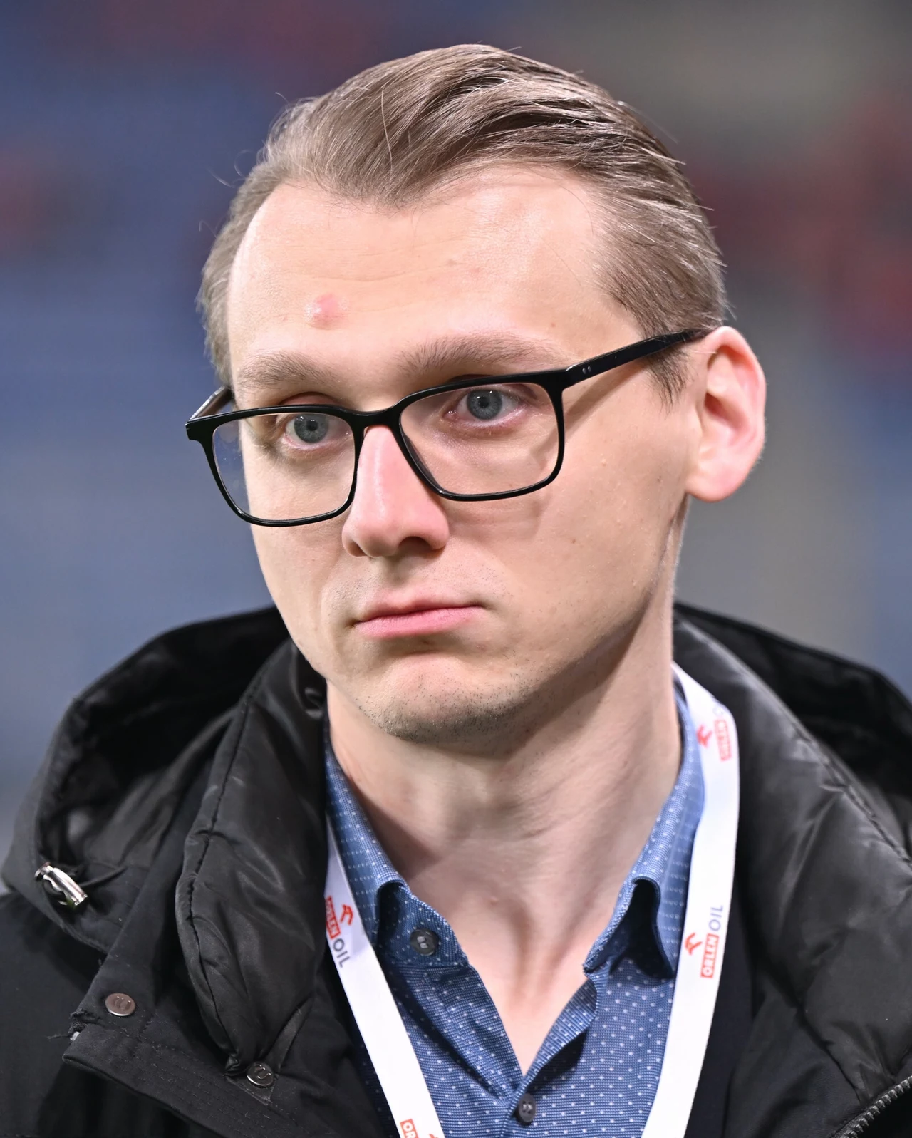 Młody mężczyzna w okularach, ubrany w niebieską koszulę i czarną kurtkę, z identyfikatorem na szyi, patrzący poważnie w stronę obiektywu, neutralne rozmyte tło.