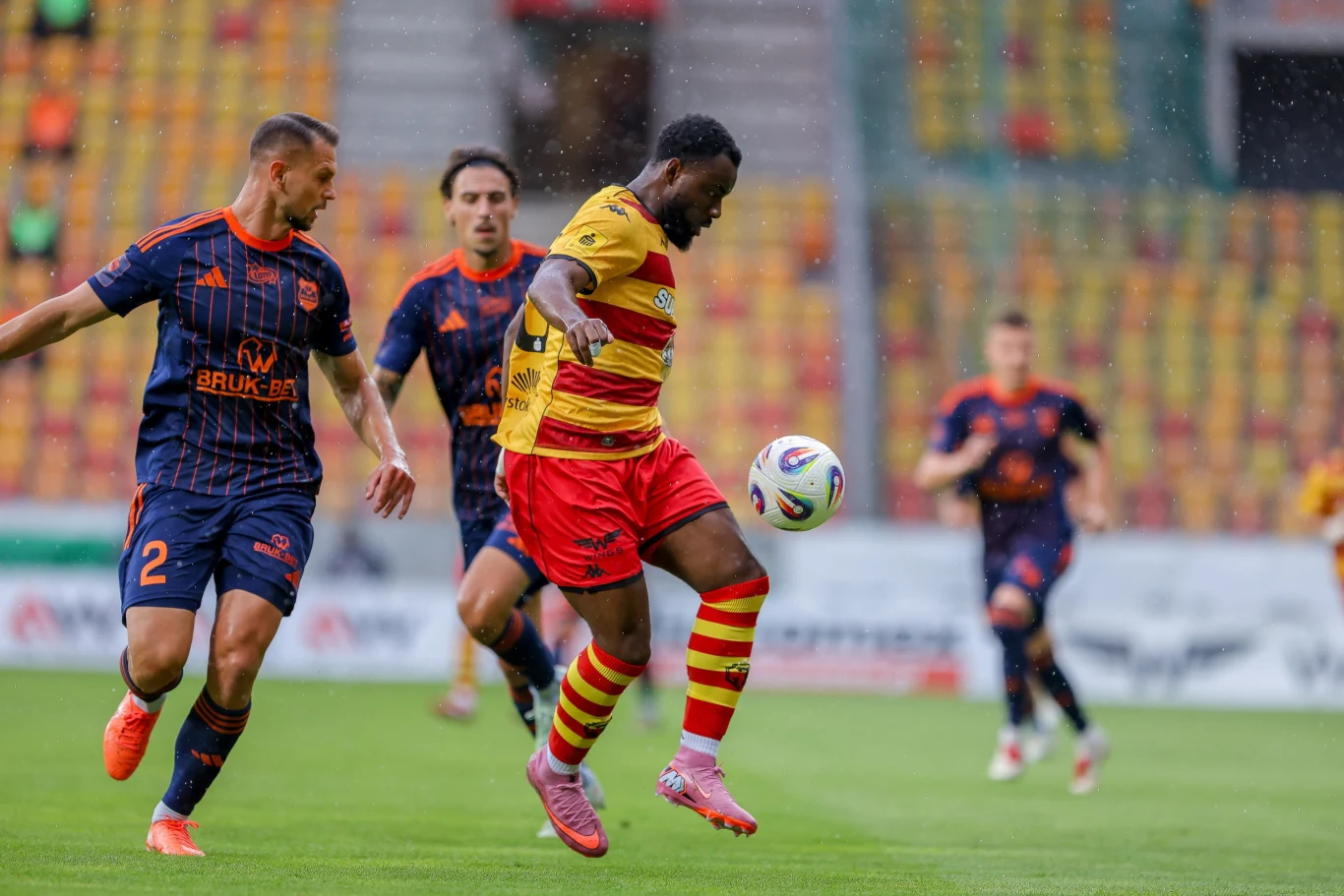 Jagiellonia Białystok - Termalica Nieciecza Piłkarz w żółto-czerwonym stroju kontroluje piłkę na boisku podczas meczu, otoczony przez zawodników drużyny przeciwnej w granatowych koszulkach, na tle pustych trybun stadionu.