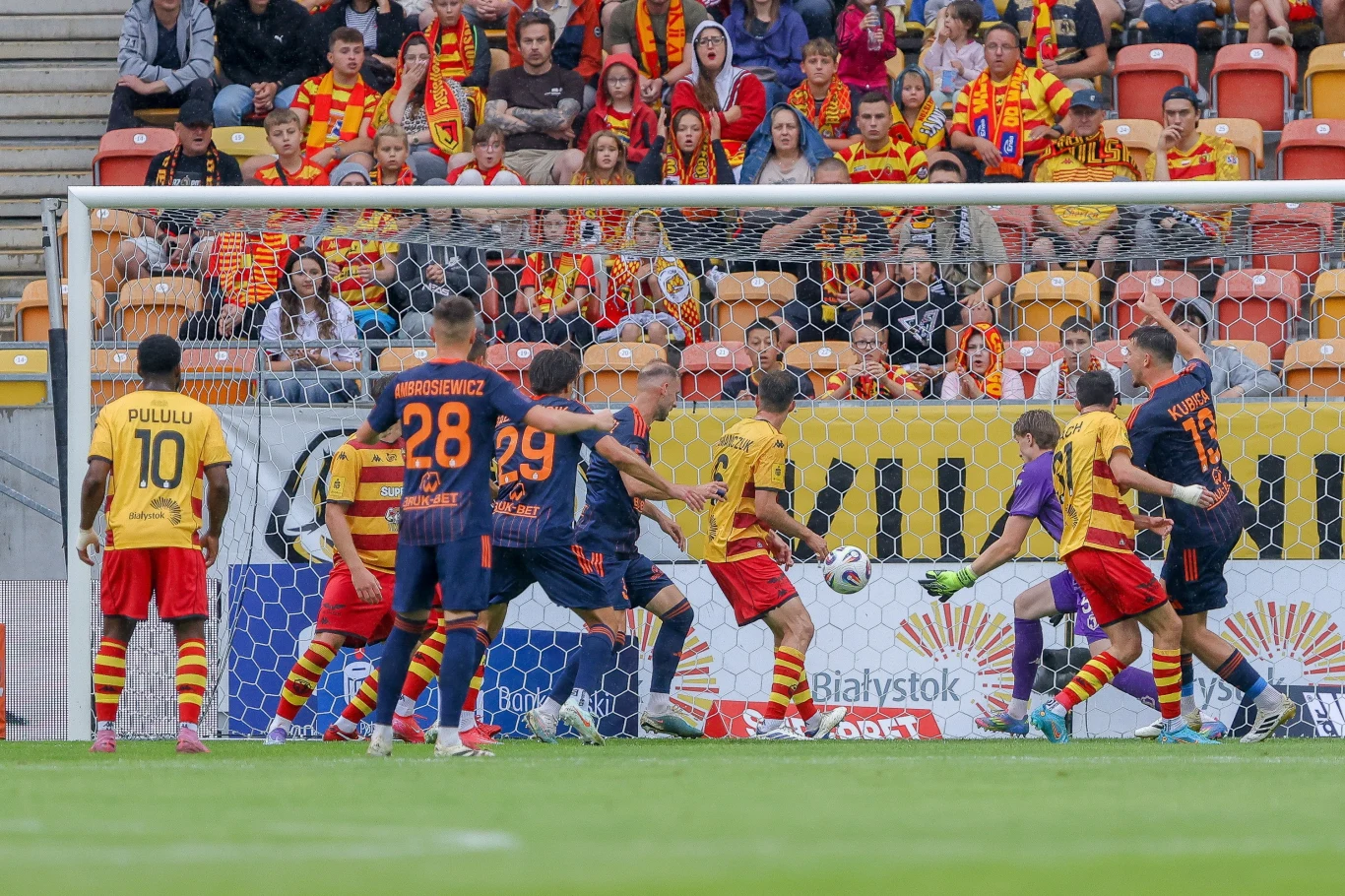 Jagiellonia Białystok - Termalica Nieciecza Intensywna akcja podczas meczu piłkarskiego, zawodnicy jednej drużyny w żółto-czerwonych strojach atakują bramkę przeciwników ubranych w granatowo-pomarańczowe koszulki, piłka znajduje się tuż przy bramce, kibice uważnie obserwują wydarzenia z trybun.