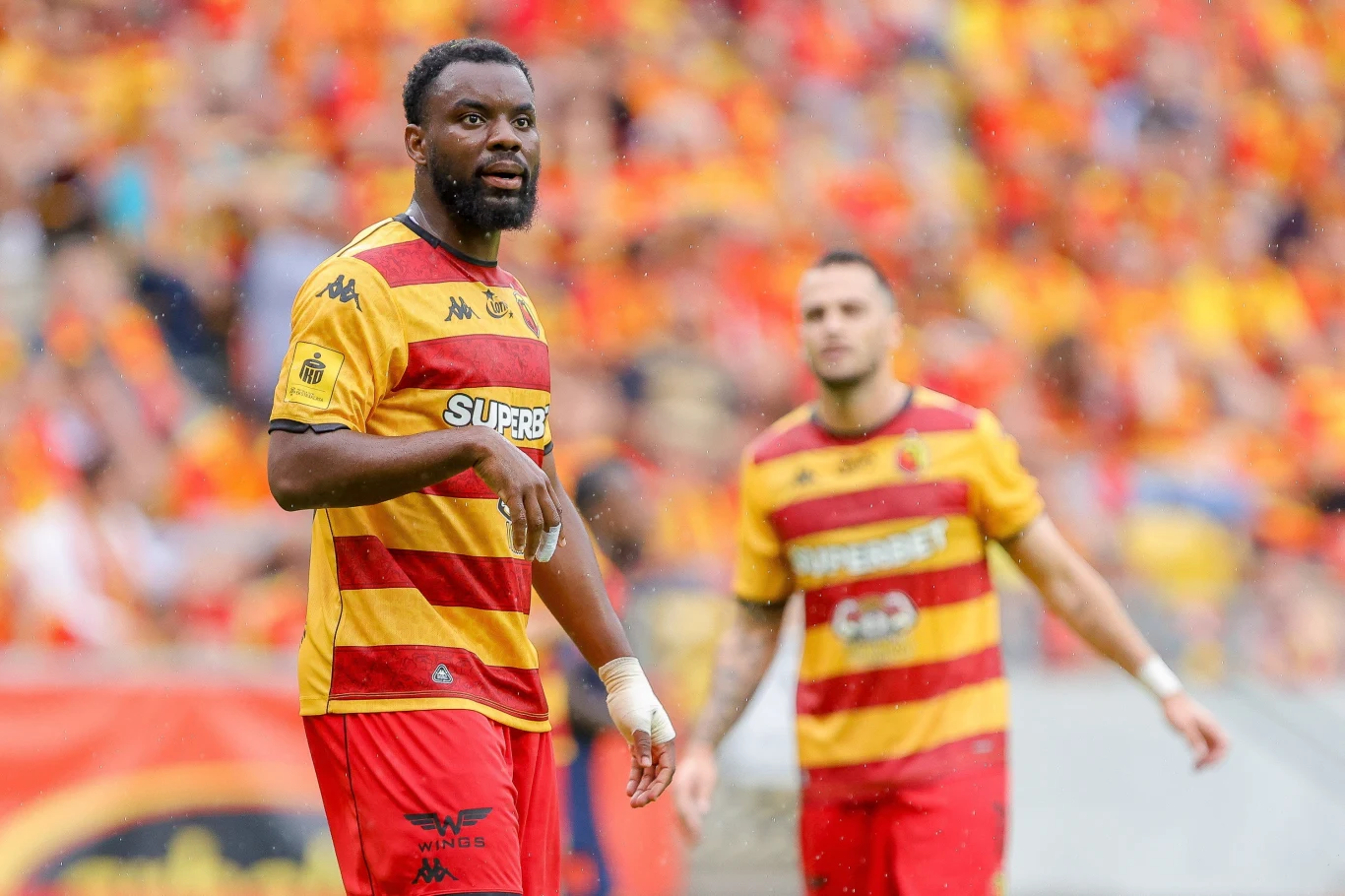 Jagiellonia Białystok - Termalica Nieciecza Dwóch piłkarzy w żółto-czerwonych strojach drużyny Jagiellonia Białystok na tle stadionu pełnego kibiców, pierwszy z nich skupiony, drugi lekko rozmyty w tle.