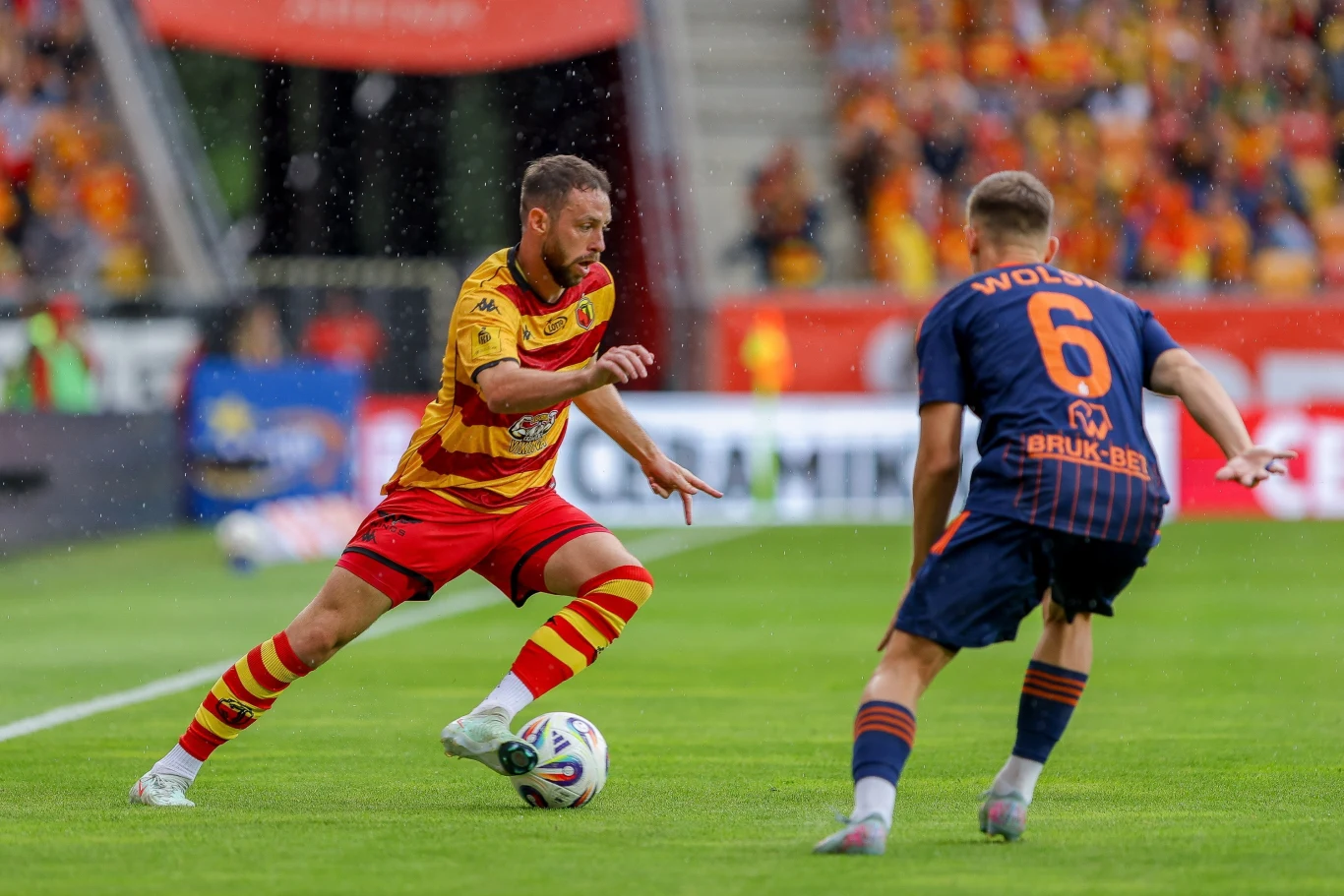 Jagiellonia Białystok - Termalica Nieciecza Dwóch piłkarzy podczas meczu piłki nożnej, zawodnik w żółto-czerwonym stroju próbuje minąć obrońcę w granatowej koszulce, na murawie stadionu w otoczeniu publiczności.