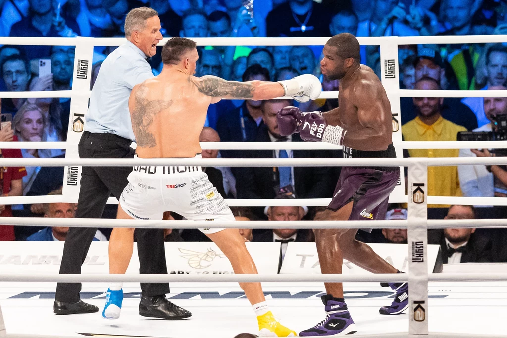 Pierwsza walka Usyk - Dubois Dwóch bokserów w trakcie wymiany ciosów na ringu, wokół nich tłum kibiców, a walkę sędziuje arbiter w niebieskiej koszuli.